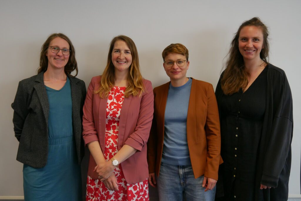 Das Foto zeigt vier Frauen, die nebeneinander vor einer weißen Wand stehen. Alle lächeln.