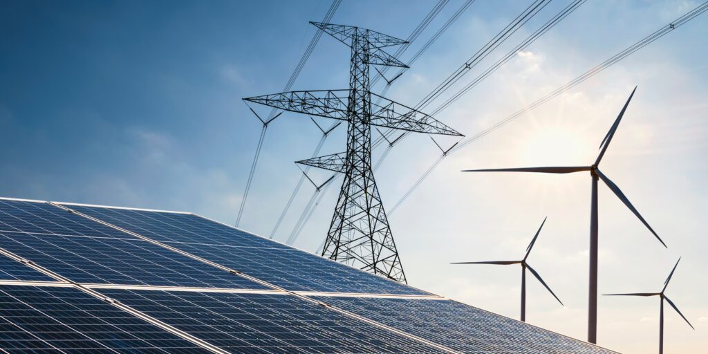 Das Bild zeigt im Vordergrund eine PV-Anlage, dahinter sind ein Strommast und Windkrafträder zu sehen.