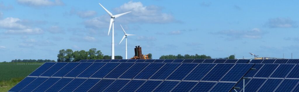 Das Foto zeigt PV-Anlagen auf einem Feld. Im Hintergrund sind zwei Windkrafträder zu sehen.