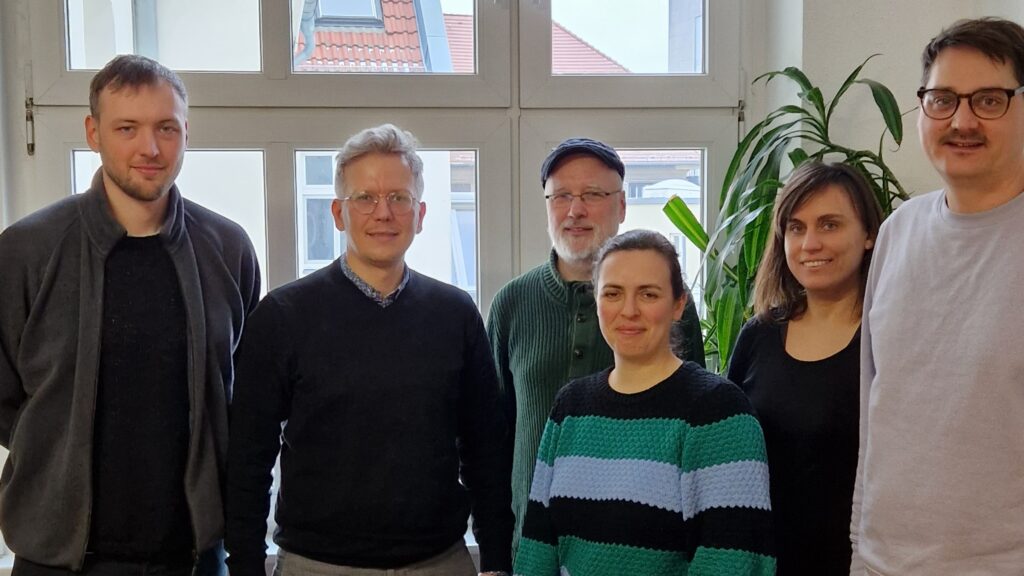 Das Foto zeigt eine Gruppe von 6 Menschen, die nebeneinander stehen und freundlich in die Kamera blicken.